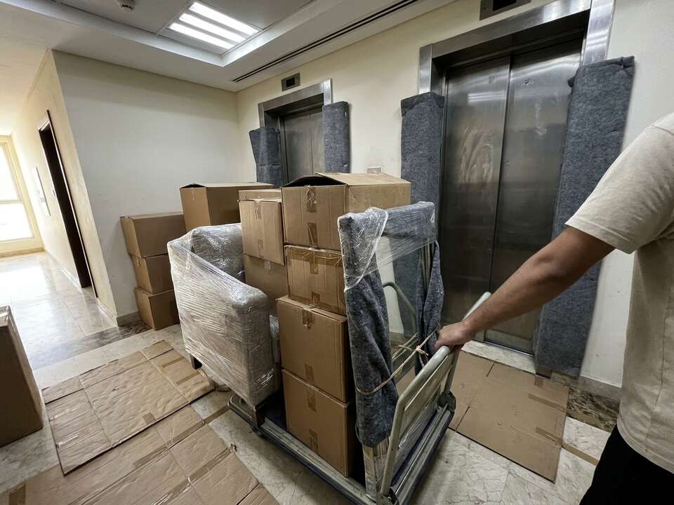 Cartons, a wrapped armchair and a padded mirror on a trolley, pushed over a protected lobby floor to padded lift doors.