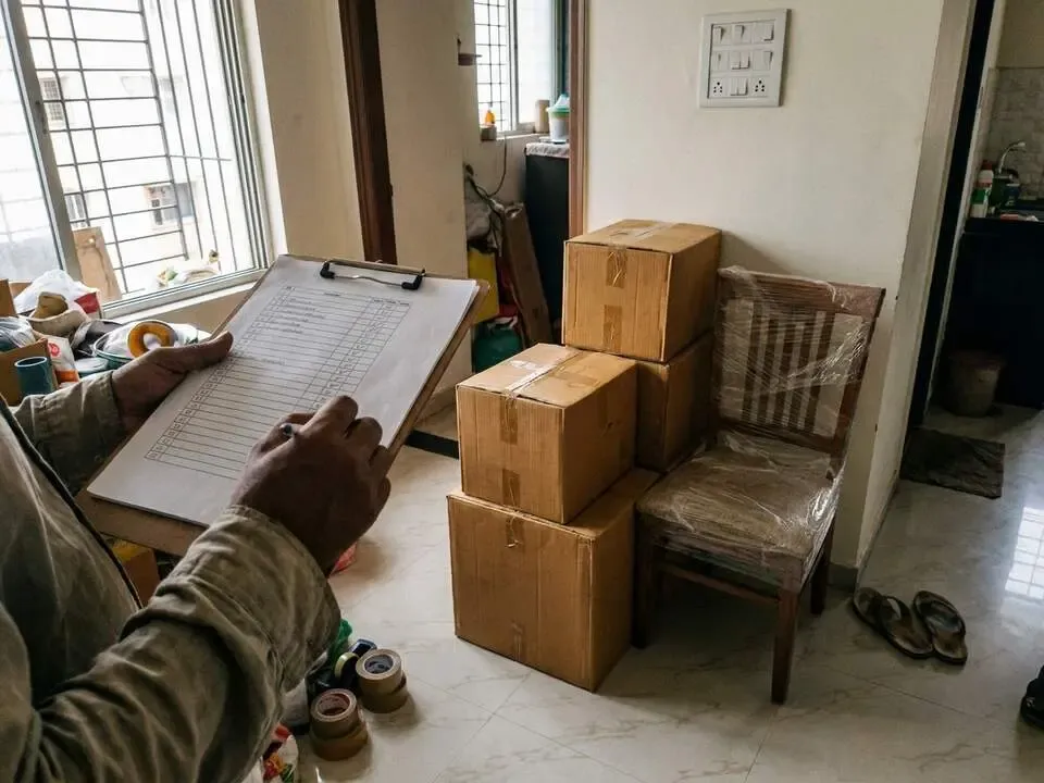 A packer ticks off an inventory on a clipboard beside taped cartons and a wrapped chair in a flat.