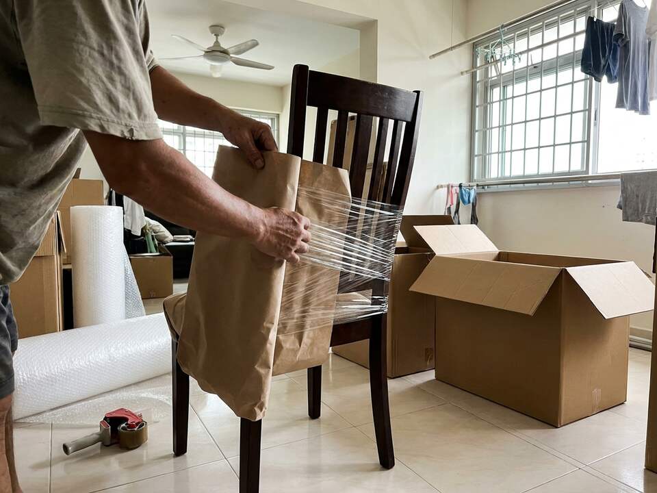 Hands wrap a wooden dining chair in brown paper and stretch film in a flat, with open cartons and bubble wrap on the floor.